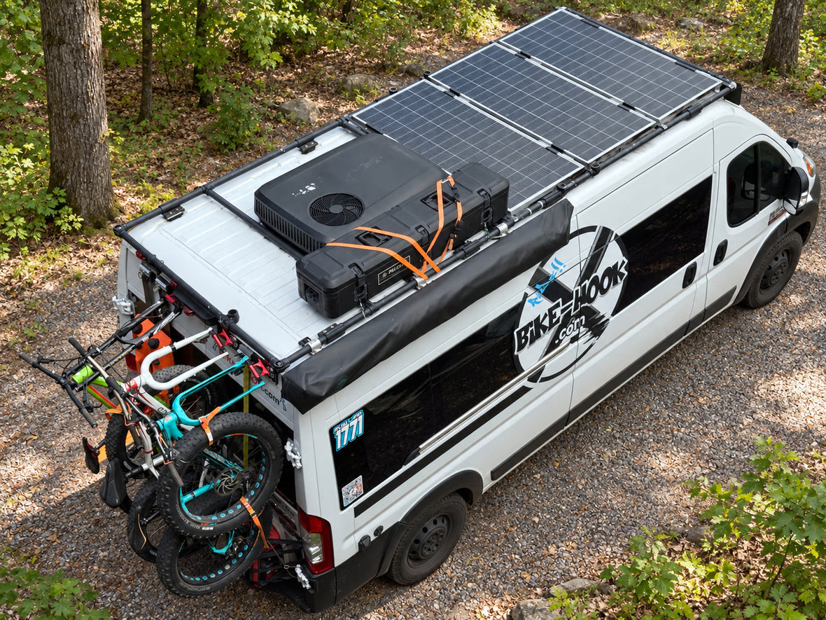 Craig’s white ProMaster with rooftop equipment and bicycles, shown against an AI-edited wooded campsite background.