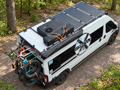 Craig’s white ProMaster with rooftop equipment and bicycles, shown against an AI-edited wooded campsite background.